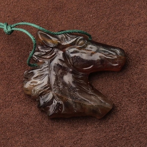 May include: A brown, translucent horse head pendant carved from a stone. The horse's mane and facial features are detailed. A green cord is attached for wearing. The pendant is displayed on a brown surface.