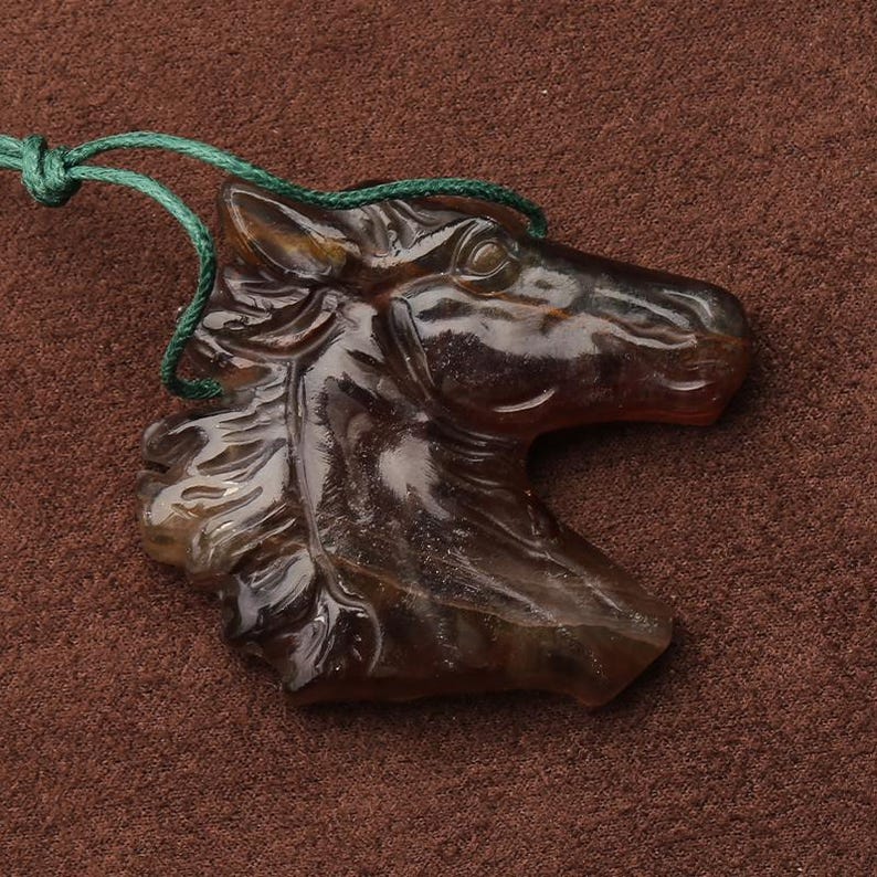 May include: A brown, translucent horse head pendant carved from a stone. The horse's mane and facial features are detailed. A green cord is attached for wearing. The pendant is displayed on a brown surface.