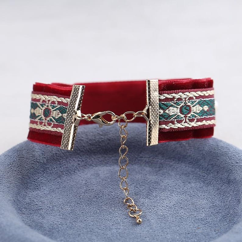 May include: A red velvet choker necklace with gold-colored clasps and a decorative embroidered band in green, white, and burgundy. The necklace has an adjustable chain for sizing.