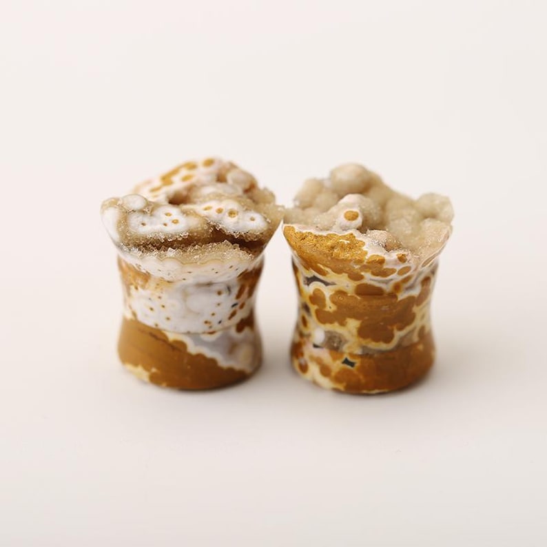 May include: Two natural stone plugs with a unique, organic appearance. The plugs feature a cylindrical shape with a textured surface. The color palette includes shades of brown, white, and tan, creating a marbled effect. The top of each plug has a crystalline formation.