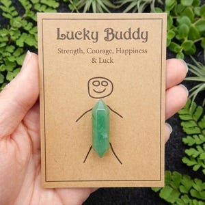 May include: A green crystal pendant in the shape of a person is attached to a brown card. The card reads "Lucky Buddy" with the words "Strength, Courage, Happiness & Luck" below. The background is a mix of green plants.