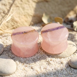 Puede incluir: Un par de dilatadores circulares para oreja hechos de piedra translúcida de color rosa claro. Cada pieza presenta una superficie lisa y pulida con una veta mineral horizontal de color púrpura oscuro que cruza el centro de la joya.