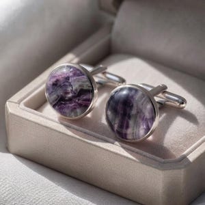Puede incluir: Un par de gemelos redondos con monturas de metal plateado pulido e incrustaciones circulares lisas de piedra veteada en color púrpura y blanco. Los gemelos se muestran uno al lado del otro, destacando las vetas naturales.