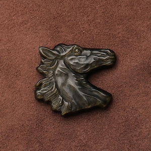 May include: A bronze-colored horse head sculpture with detailed mane and facial features. The sculpture is set against a brown background, highlighting the intricate design and texture of the horse's head.