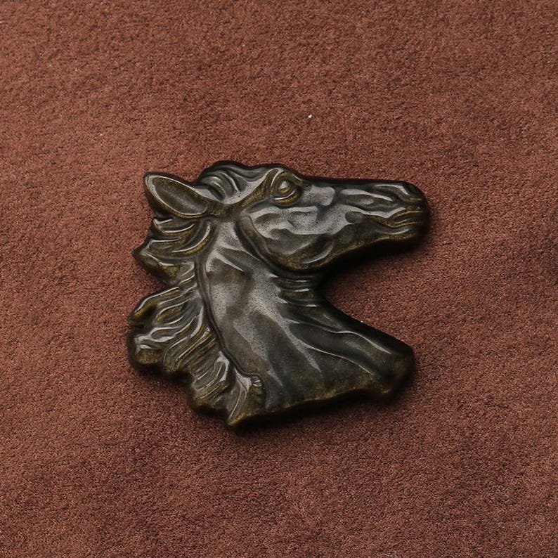 May include: A bronze-colored horse head sculpture with detailed mane and facial features. The sculpture is set against a brown background, highlighting the intricate design and texture of the horse's head.