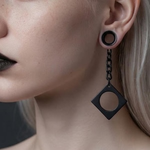 May include: A matte black ear plug featuring a circular tunnel design connected to a short chain link that suspends a square pendant with a circular cutout in the center. The jewelry is crafted from a dark metal material with a smooth finish.