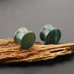 May include: A pair of cylindrical ear plugs crafted from polished green stone with natural swirling patterns. The plugs feature a smooth, glossy finish and a concave shape, showcasing deep forest green and lighter teal hues throughout the material.
