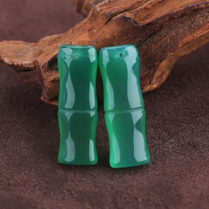 May include: Two green, bamboo-shaped beads with a glossy finish. The beads are a vibrant shade of green and have a smooth, polished surface. They are displayed against a backdrop of dark wood and a brown surface.