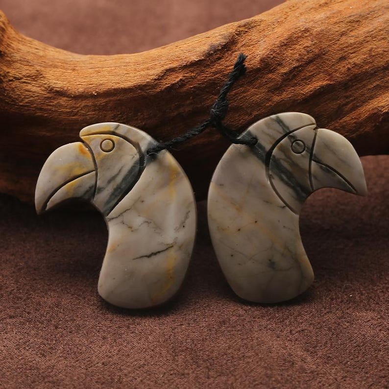 May include: Two carved stone pendants shaped like bird heads. The pendants are connected by a black string. The stone has a marbled appearance with shades of gray, cream, and yellow. The bird heads have detailed beaks and eye sockets.
