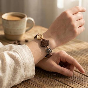 May include: A brown bracelet with square and button-shaped wooden beads and small metal charms. The bracelet is worn on a wrist, with a cup of coffee in the background. The bracelet is on a wooden surface.