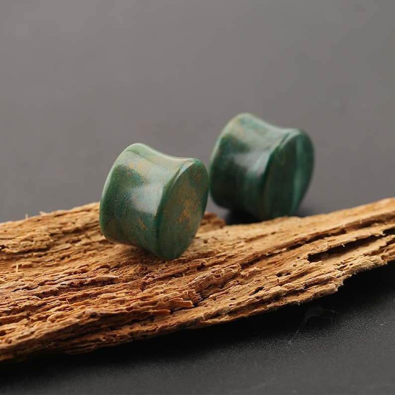May include: A pair of cylindrical ear plugs with a smooth, polished finish. The plugs feature a deep forest green color with subtle, swirling golden-brown mineral inclusions throughout the surface, creating a natural, stone-like appearance.