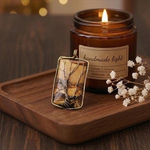 May include: A rectangular pendant with a tree design, set in a gold-colored frame, rests on a wooden tray. A lit candle in a brown glass jar is in the background. The candle has a label that reads "handmade light".