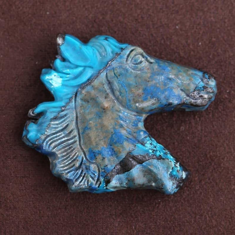 May include: A carved turquoise horse head sculpture. The sculpture features a detailed mane and facial features. The horse head is primarily turquoise blue with areas of darker blue and gray. The sculpture is set against a brown background.