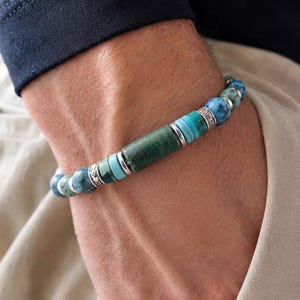 Può includere: Un braccialetto di perline con una perla centrale cilindrica in pietra verde, affiancata da perline a disco più piccole color turchese, perline rotonde in pietra screziata blu-verde e distanziatori in metallo argentato con motivi incisi.