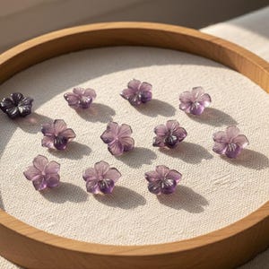 May include: A collection of small, translucent purple flower-shaped beads arranged on a light-colored fabric within a wooden embroidery hoop. The beads have detailed petal designs and are evenly spaced.