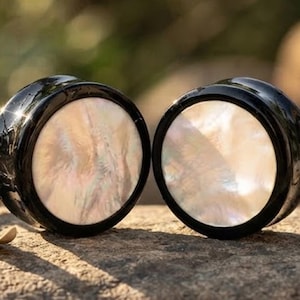 May include: A pair of circular ear plugs featuring a smooth, glossy black outer rim and a flat, iridescent white mother-of-pearl inlay. The center of each plug displays a swirling, pearlescent texture that reflects light with subtle pink and blue tones.
