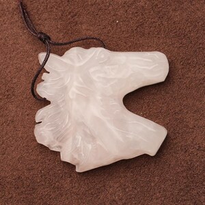 May include: A white, translucent horse head pendant carved from a stone, with a dark brown cord attached. The pendant is detailed with a mane and facial features. The background is a solid brown.