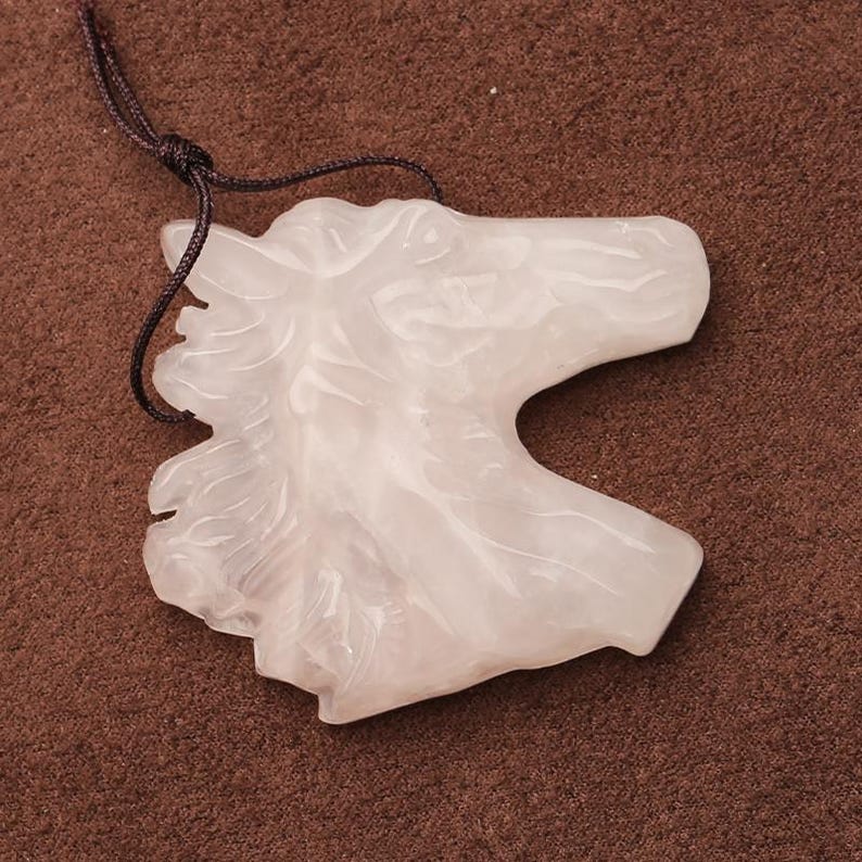 May include: A white, translucent horse head pendant carved from a stone, with a dark brown cord attached. The pendant is detailed with a mane and facial features. The background is a solid brown.