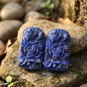 May include: Two rectangular deep blue lapis lazuli pendants featuring intricate relief carvings of blooming flowers and foliage. Each pendant has a small drilled hole at the top for suspension, showcasing a smooth, polished surface texture.