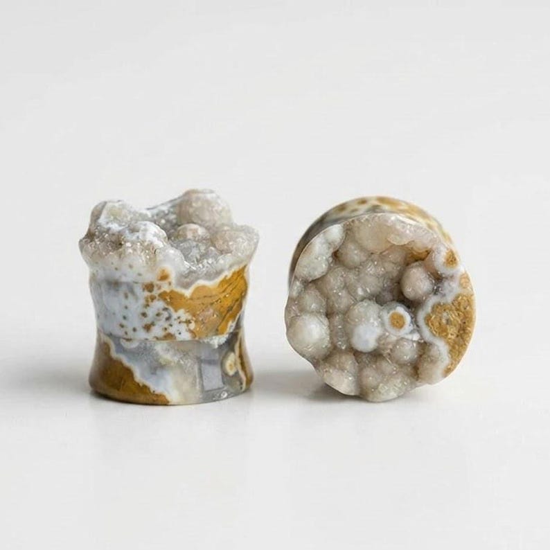 May include: Two stone plugs with a unique, natural appearance. One plug is cylindrical, the other is a rounded shape. The plugs feature a mix of colors, including white, tan, and brown, with a textured, crystal-like surface.