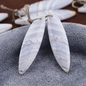 May include: Two blue lace agate pendants with a light blue and white banded pattern. Each pendant is elongated with a rounded tip and a small hole at the top for stringing. The pendants are displayed on a grey cloth.