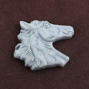 May include: A light blue horse head sculpture with detailed mane and facial features. The sculpture is made of a smooth material and is set against a brown background. The horse head is a decorative item.