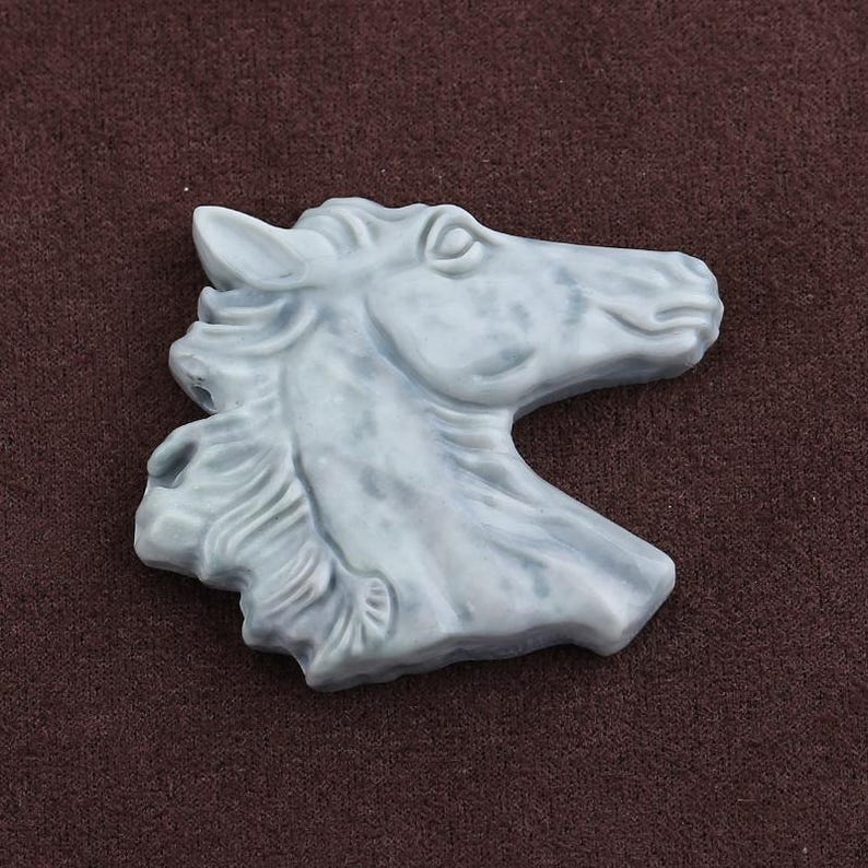 May include: A light blue horse head sculpture with detailed mane and facial features. The sculpture is made of a smooth material and is set against a brown background. The horse head is a decorative item.