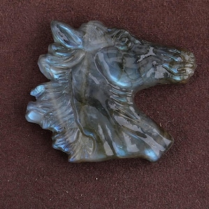 May include: A carved stone sculpture of a horse's head. The sculpture is a mix of gray and blue hues, with iridescent flashes. The horse's mane and facial features are detailed. The sculpture is set against a brown background.