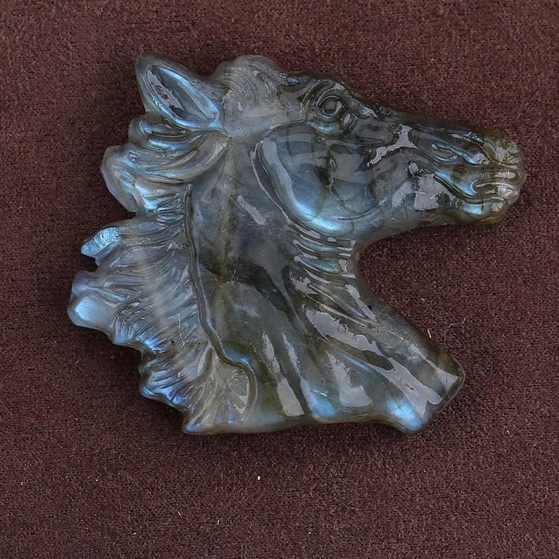 May include: A carved stone sculpture of a horse's head. The sculpture is a mix of gray and blue hues, with iridescent flashes. The horse's mane and facial features are detailed. The sculpture is set against a brown background.
