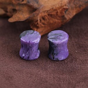 May include: A pair of cylindrical ear plugs crafted from polished purple charoite stone, featuring a smooth concave waist and natural swirling black and white mineral inclusions throughout the vibrant violet surface of each piece.