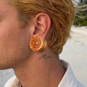 May include: A circular ear plug featuring a detailed, golden-brown ammonite fossil pattern with a spiral design. The plug is inserted into a stretched earlobe, and a small, simple black line tattoo of a wave is visible on the neck below the ear.