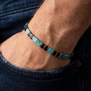 May include: A beaded bracelet featuring alternating square-shaped segments of turquoise-coloured stone with dark veining and smooth black beads, separated by thin metallic gold-toned spacers, arranged in a repeating pattern around the wrist.