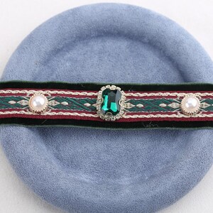Pode incluir: Um colar gargantilha decorativo com uma faixa de veludo verde e um design estampado em vermelho e verde. A gargantilha apresenta uma grande gema verde esmeralda e dois enfeites perolados, sobre um fundo azul claro.