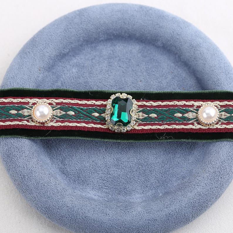 Pode incluir: Um colar gargantilha decorativo com uma faixa de veludo verde e um design estampado em vermelho e verde. A gargantilha apresenta uma grande gema verde esmeralda e dois enfeites perolados, sobre um fundo azul claro.