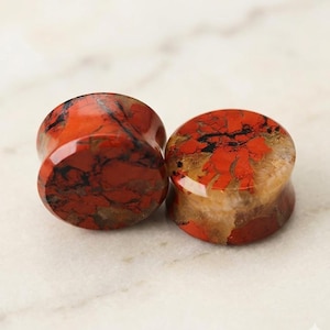 May include: Two red jasper stone ear plugs with black veining. The plugs have a flared design and a polished finish. The colors range from deep red to light tan, with black streaks throughout. The plugs are displayed on a white surface.