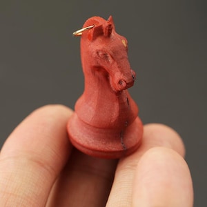 May include: A red wooden chess knight piece with a gold-colored ring attached at the top. The knight is carved in detail, showing the horse's head and neck. The piece is held in a hand against a gray background.