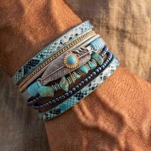 May include: A multi-strand bracelet featuring light blue snakeskin-patterned leather, a silver-toned metal feather charm with a central turquoise stone, small turquoise beads, and dark blue spherical beads arranged in a layered, stacked design.
