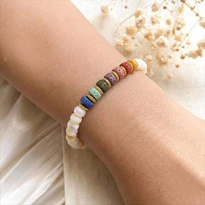 May include: A colorful beaded bracelet featuring a mix of rainbow-colored beads and white pearl-like beads. The bracelet is accented with gold-colored spacers. The bracelet is worn on a wrist.