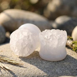 May include: A pair of circular ear plugs featuring a smooth, translucent white body and a textured, crystalline front surface. The plugs have a concave profile, narrowing slightly in the centre, with a jagged, mineral-like appearance on the face.