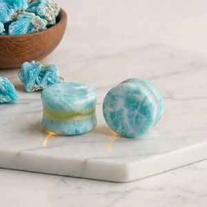 May include: Two light blue and white stone plugs with a yellow band are displayed on a white marble surface. A wooden bowl filled with similar blue stones sits in the background, along with loose stones.