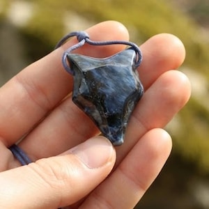 Può includere: Un ciondolo intagliato a forma di testa di volpe in una pietra blu scuro e nera con finitura lucida. Il ciondolo è sospeso a un cordino sottile azzurro che passa attraverso la parte superiore dell'intaglio in pietra per formare una collana.