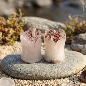 May include: A pair of ear plugs crafted from pale pink quartz with a concave cylindrical shape. The top edges feature jagged, crystalline points with natural reddish-brown mineral inclusions embedded within the translucent stone structure.