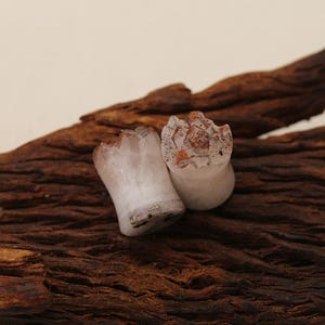 May include: Two ear plugs with a cylindrical shape and a flared base. The body is a translucent off-white material with dark speckles. The top surface features a jagged, crystalline texture with reddish-brown and orange mineral inclusions.