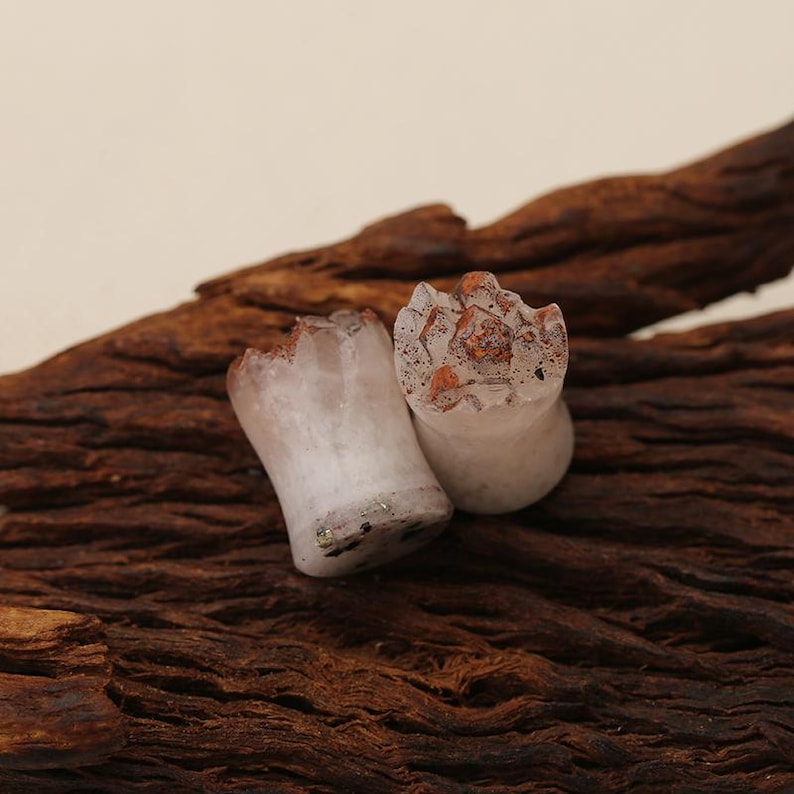 May include: Two ear plugs with a cylindrical shape and a flared base. The body is a translucent off-white material with dark speckles. The top surface features a jagged, crystalline texture with reddish-brown and orange mineral inclusions.