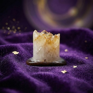 Puede incluir: Un portavelas cilíndrico de cristal de citrino con una base marrón oscuro. La parte superior del cristal es un grupo de puntas de color amarillo dorado. El portavelas se encuentra sobre una superficie de terciopelo morado intenso con detalles de pan de oro.