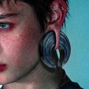 May include: A crescent-shaped ear gauge plug with a dark blue and black marbled finish. The jewelry features a smooth, curved design that fits through a stretched earlobe, complemented by a small silver-toned hoop earring in the upper ear cartilage.