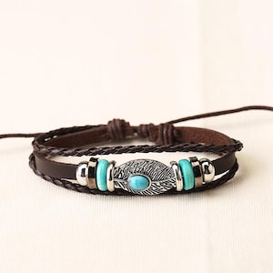 Könnte beinhalten: Braunes Lederarmband mit geflochtenem Band und zentralem Dekorationselement. Das Mittelstück zeigt ein silbernes Federdesign mit einem türkisfarbenen Stein und türkisfarbenen Perlen. Das Armband hat einen verstellbaren Verschluss.