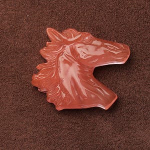 May include: A reddish-orange horse head sculpture. The horse head is carved with detailed features, including the mane, eye, and muzzle. The sculpture is made of a translucent material and is set against a brown background.