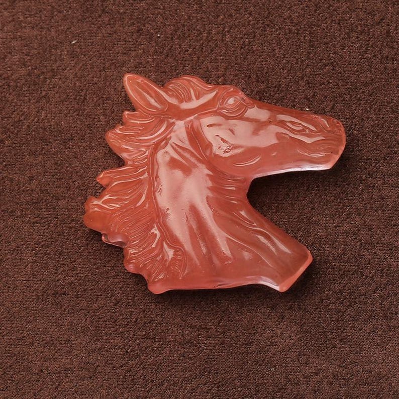 May include: A reddish-orange horse head sculpture. The horse head is carved with detailed features, including the mane, eye, and muzzle. The sculpture is made of a translucent material and is set against a brown background.