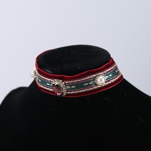 May include: A burgundy velvet choker necklace with a green and gold patterned ribbon. The choker features a large, oval, purple gemstone and two pearls. The necklace is displayed on a black mannequin head.